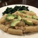 pasta with white bean sauce topped with sliced avocado with a bed of kale in the background
